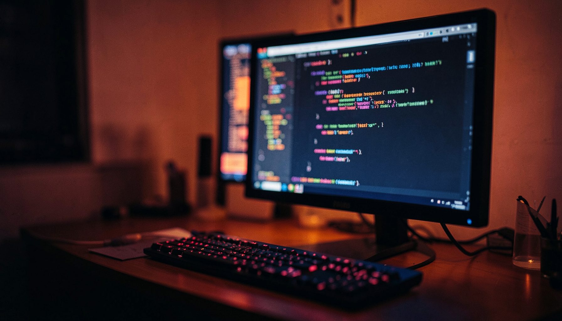 A wide cinematic frame of a dimly lit desk with a glowing monitor displaying colorful code syntax, warm amber and deep blue tones, shallow depth of field
